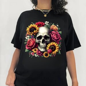 Puede incluir: Una camiseta negra con un gr&aacute;fico central de un cr&aacute;neo humano rodeado por un vibrante arreglo de flores coloridas, que incluye girasoles amarillos, rosas rojas y flores naranjas, creando un dise&ntilde;o floral detallado y contrastante.