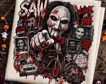 May include: A white t-shirt featuring a graphic print of the Saw movie character Billy the Puppet pointing forward. The design includes text reading SAW, LIVE OR DIE, LET THE GAME BEGIN, MAKE YOUR CHOICE, PLAY ME, and ALL PUZZLES HAVE A SOLUTION.