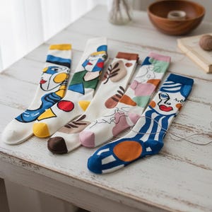 May include: Five pairs of white socks with colorful abstract face designs. Each sock features a different design with various colors including blue, yellow, red, and brown. The socks are arranged on a light-colored wooden surface.