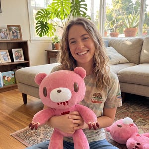May include: A pink plush bear with brown claws and a bloody mouth is held by a person. The bear has a white muzzle and brown ears. A second bear is on the floor. The person is wearing a tie-dye shirt with a California bear graphic.