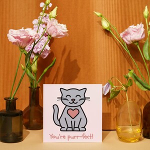 Puede incluir: Una tarjeta de felicitación con una ilustración de un gato gris y un corazón rosa, con el texto "You're purr-fect!". La tarjeta está rodeada de jarrones con flores rosas, creando una composición encantadora.