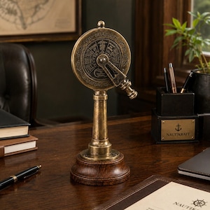 May include: A brass ship telegraph model with a wooden base. The circular dial has engraved text and a central handle. The base is dark brown, and the telegraph is displayed on a wooden desk. The dial reads "SHIP TELEGRAPH" and "LIVERPOOL, LON".