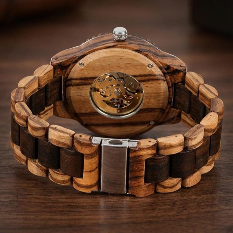 May include: Wooden wristwatch with a visible mechanical movement and a wooden strap. The watch case and strap feature a striped wood grain pattern in shades of brown and tan. The watch has a silver-coloured crown and clasp.
