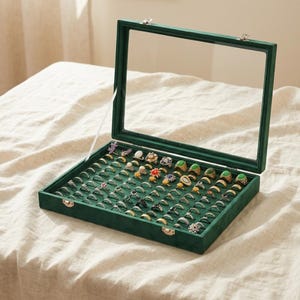 Puede incluir: Un joyero rectangular de terciopelo verde oscuro con tapa de cristal transparente, que cuenta con múltiples ranuras organizadas en filas para albergar una gran colección de anillos de metal con diversos diseños y gemas.