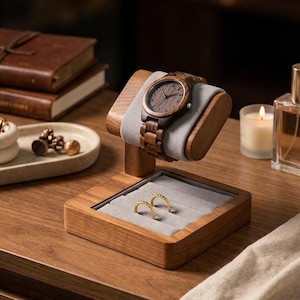 May include: A wooden watch with a dark brown face and band rests on a wooden stand with a gray cushion. Below, a wooden tray holds two gold rings. The watch stand is on a wooden surface.