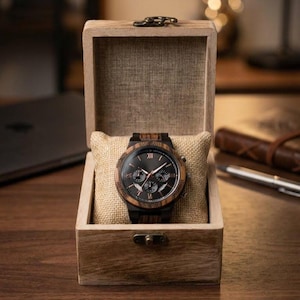 Peut inclure: Une montre-bracelet en bois avec un cadran noir et des accents bruns repose sur un coussin beige à l'intérieur d'une boîte en bois. La montre comporte des chiffres romains et trois sous-cadrans. La boîte a un couvercle à charnière et un petit fermoir.