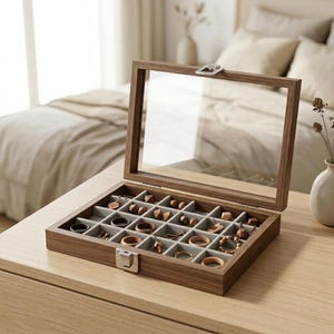 Puede incluir: Una caja de joyería de madera con tapa de cristal transparente, abierta para exhibir varios anillos y pendientes. La caja está dividida en compartimentos con forro de tela gris. Las joyas son de madera y metal.