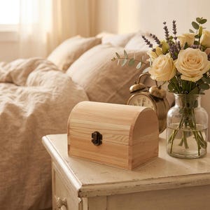 May include: A light-coloured wooden chest with a curved lid and a small metal latch on the front. The box is constructed from natural pine wood with visible grain patterns and is positioned on a white surface next to a glass vase of flowers.