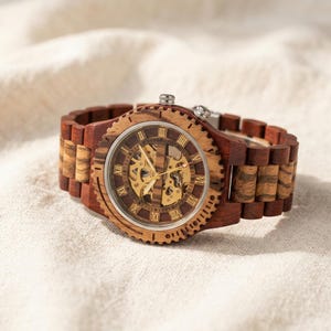 May include: A wristwatch featuring a dark and light brown wood link band and a circular wood bezel. The watch face displays a skeletonized gold-tone mechanical movement, gold-tone hands, and gold-tone Roman numeral hour markers.