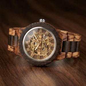 May include: A wooden wristwatch with a visible mechanical movement. The watch has a dark brown bezel and a light brown wooden strap. The watch face features Roman numerals and gold-coloured gears.
