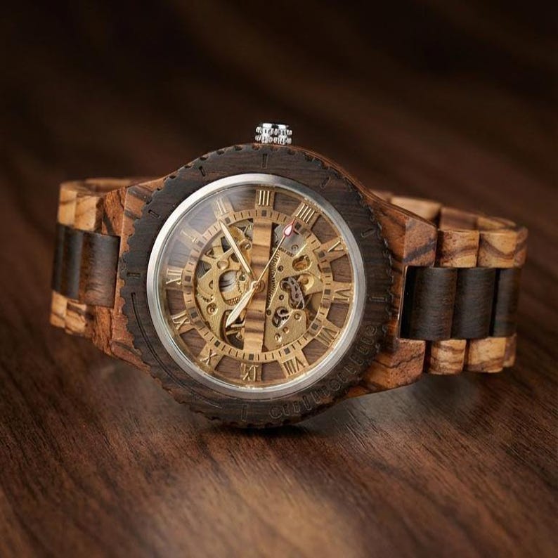 May include: A wooden wristwatch with a visible mechanical movement. The watch has a dark brown bezel and a light brown wooden strap. The watch face features Roman numerals and gold-coloured gears.