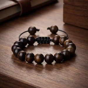 May include: A dark brown wooden bead bracelet. The bracelet features multiple round beads strung together on a black cord. The beads have a natural wood grain pattern. The bracelet is displayed on a wooden surface.