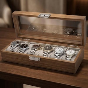 Peut inclure: Un coffret en bois pour montres avec un couvercle en verre transparent, présentant six montres-bracelets avec des bracelets argentés et dorés. Le coffret est ouvert, révélant les montres sur des coussins crème.
