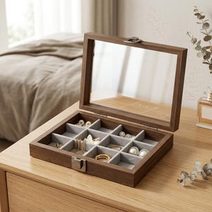 Puede incluir: Una caja de joyería de madera con tapa de cristal transparente, abierta para exhibir varias joyas. La caja está dividida en compartimentos con forro de tela gris. Las joyas incluyen collares, anillos y pendientes. La caja está sobre una superficie de madera.