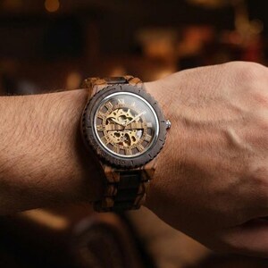 May include: A wooden wristwatch with a visible mechanical movement. The watch has a dark brown wooden bezel and band, with a clear glass face displaying gold-coloured gears and Roman numeral hour markers. The watch is on a wrist.