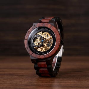 Peut inclure: Une montre-bracelet avec un boîtier rond en bois et un bracelet à maillons alternant des segments de bois brun foncé et brun rougeâtre. Le cadran affiche le texte BOBO BIRD, un mouvement mécanique doré squeletté et un petit sous-cadran.