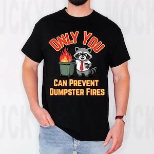 Op de afbeelding: Zwart t-shirt met een cartoon wasbeer met een stropdas die een koffiekopje vasthoudt, naast een brandende vuilnisbak. De tekst luidt "ONLY YOU CAN PREVENT DUMPSTER FIRES" in rood en geel.