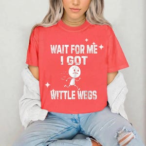 Puede incluir: Camiseta roja con el texto blanco "WAIT FOR ME I GOT WITTLE WEGS". La camiseta presenta un muñeco de palo corriendo con una cara triste. La camiseta la lleva una persona con vaqueros azules.