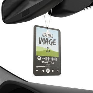 May include: Rectangular car air freshener with the text "UPLOAD IMAGE" above a landscape graphic. Below is a music player interface with song details. The air freshener hangs from a string, suspended in front of a car's rearview mirror.