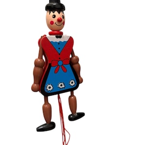 May include: A wooden pull-string puppet with a painted face, black hat, and a red and blue outfit. The puppet has jointed arms and legs, and a red string hangs down with a red ball at the end. The puppet's design is reminiscent of a traditional toy.