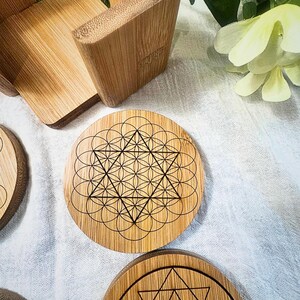 May include: A set of round bamboo coasters with intricate geometric designs etched in black. The coasters feature patterns like the Flower of Life and other sacred geometry symbols. A bamboo coaster holder is also visible.