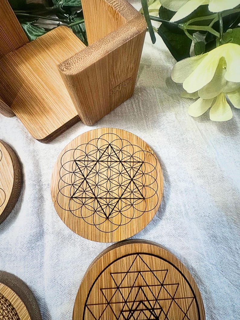 May include: A set of round bamboo coasters with intricate geometric designs etched in black. The coasters feature patterns like the Flower of Life and other sacred geometry symbols. A bamboo coaster holder is also visible.