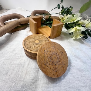 May include: A set of four round bamboo coasters with intricate geometric designs. One coaster features a Metatron's Cube pattern, while another displays a radial design. The coasters are stacked next to a bamboo holder.