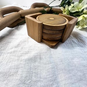 May include: A set of bamboo coasters with a geometric design, held in a matching square holder. The coasters are stacked, with the top one showing a detailed spiral pattern. The holder has a natural wood grain finish.