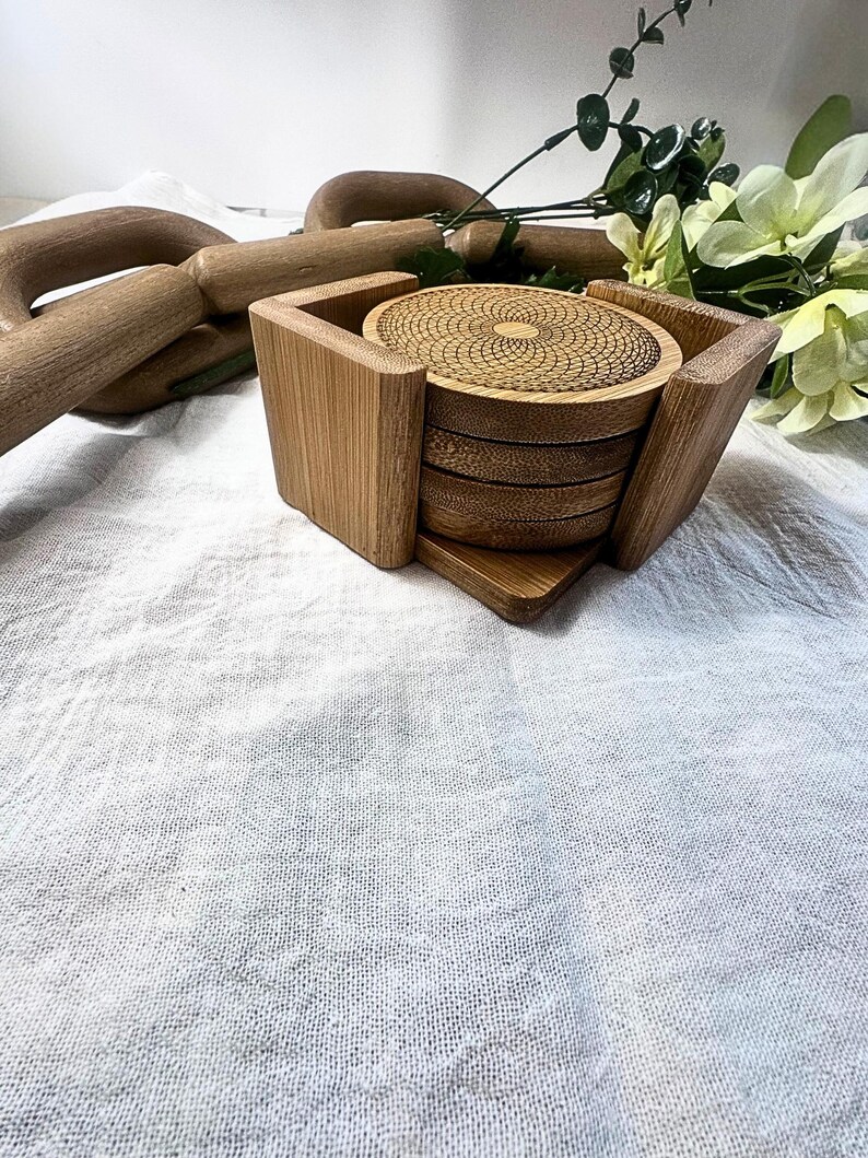 May include: A set of bamboo coasters with a geometric design, held in a matching square holder. The coasters are stacked, with the top one showing a detailed spiral pattern. The holder has a natural wood grain finish.