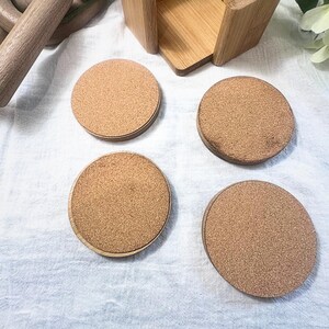 May include: A set of four round cork coasters with a light brown color, each with a wooden border. They are displayed with a wooden holder and other wooden decorative items on a white surface. The coasters are designed to protect surfaces from drink rings.