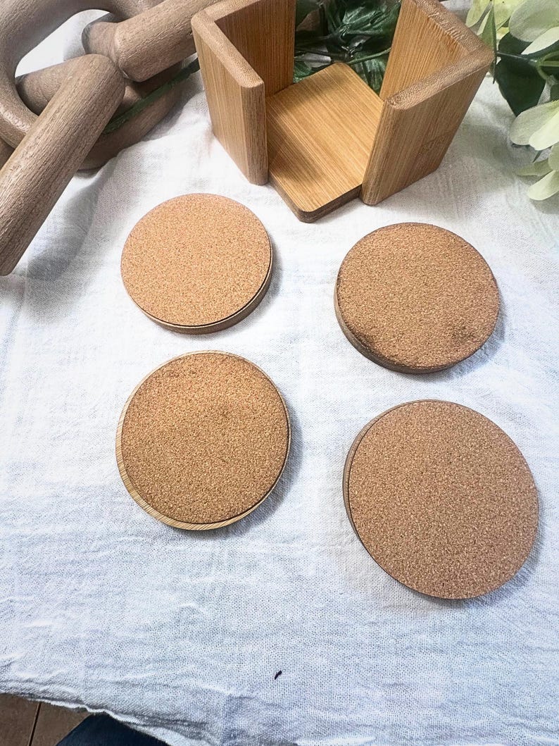May include: A set of four round cork coasters with a light brown color, each with a wooden border. They are displayed with a wooden holder and other wooden decorative items on a white surface. The coasters are designed to protect surfaces from drink rings.