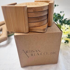 May include: A set of bamboo coasters with a holder, presented on a cardboard box. The coasters are round with a geometric design etched on top. The holder is square and made of the same light-colored bamboo.