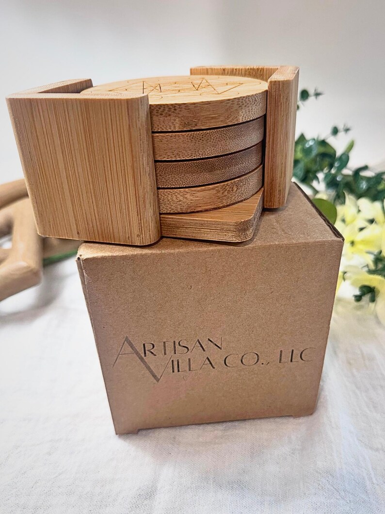 May include: A set of bamboo coasters with a holder, presented on a cardboard box. The coasters are round with a geometric design etched on top. The holder is square and made of the same light-colored bamboo.