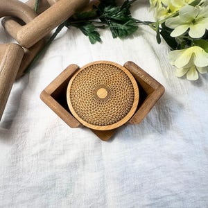 May include: A wooden coaster with a geometric design sits in a matching holder. The coaster and holder are made of light-colored wood. The coaster has a detailed, etched pattern. The background includes flowers and other wooden objects.