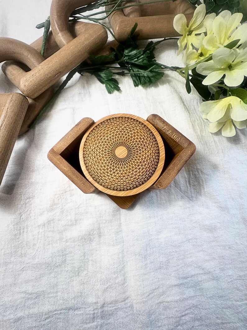 May include: A wooden coaster with a geometric design sits in a matching holder. The coaster and holder are made of light-colored wood. The coaster has a detailed, etched pattern. The background includes flowers and other wooden objects.