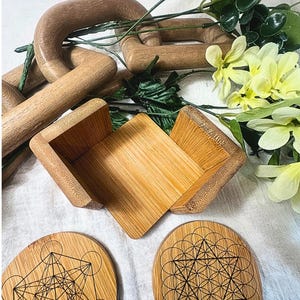 May include: A collection of wooden home decor items, including a small open box, two oval coasters with geometric designs, and a wooden chain. Greenery and yellow flowers are arranged around the items.