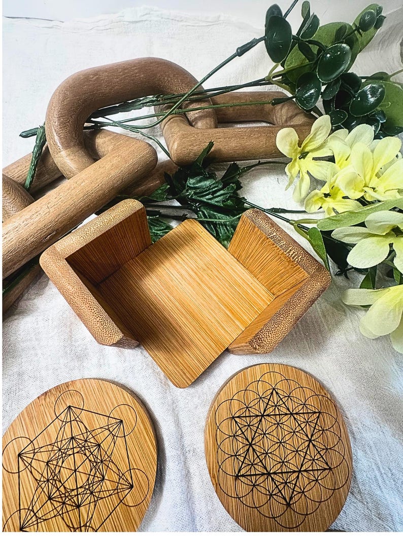 May include: A collection of wooden home decor items, including a small open box, two oval coasters with geometric designs, and a wooden chain. Greenery and yellow flowers are arranged around the items.