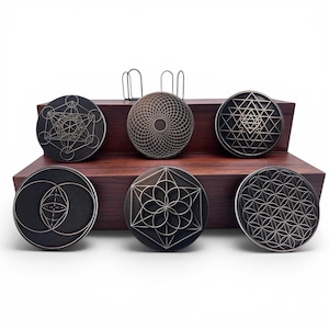 May include: A set of six circular black coasters featuring intricate metallic geometric engravings. Each coaster displays a unique pattern, including the Flower of Life, Metatron's Cube, and Sri Yantra, arranged on a two-tiered dark wood display stand.