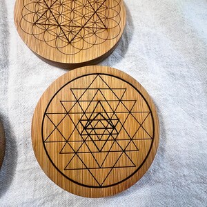 May include: Wooden coasters with geometric designs. One coaster features a Flower of Life pattern, while another displays a Sri Yantra design. The coasters are made of light brown wood and have dark brown etched patterns.