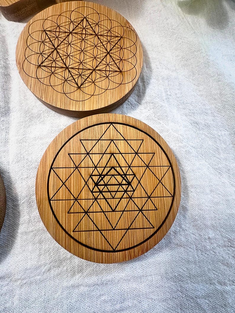 May include: Wooden coasters with geometric designs. One coaster features a Flower of Life pattern, while another displays a Sri Yantra design. The coasters are made of light brown wood and have dark brown etched patterns.