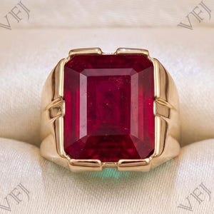 May include: A gold ring featuring a large, rectangular, emerald-cut deep red gemstone set in a polished gold band. The ring is displayed inside a dark wood jewelry box with a cream-colored interior lining. The letters VFJ are visible on the image.