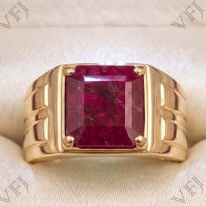 Könnte beinhalten: Ein Goldring mit einem großen, rechteckigen, tiefroten Edelstein in der Mitte. Die Ringschiene ist poliert und weist an den Seiten gerillte Details auf. Der Ring befindet sich in einer dunklen Holzschatulle mit cremefarbenem Innenfutter.
