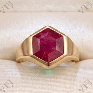 Könnte beinhalten: Ein Goldring mit einem großen, sechseckig geschliffenen, tiefroten Rubin, der in eine glatte, polierte Goldschiene gefasst ist. Der Ring wird in einer dunklen Holzschatulle mit cremefarbenem, gepolstertem Innenfutter präsentiert.