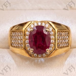 Puede incluir: Un anillo de oro con un gran rubí rojo oscuro de talla ovalada en el centro, rodeado por un halo de pequeños diamantes blancos redondos. La banda es ancha e incluye diamantes blancos adicionales engastados en pavé a ambos lados.