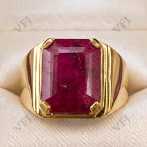 Könnte beinhalten: Ein Goldring mit einem großen, rechteckigen, tiefroten Edelstein mit sichtbaren natürlichen Einschlüssen, gefasst in eine polierte Goldschiene. Der Ring befindet sich in einer Schmuckschatulle aus dunklem Holz mit cremefarbenem Satinfutter.