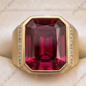 May include: A gold ring featuring a large, rectangular, deep red gemstone set in the center. The band is wide and smooth, accented with a vertical row of small, clear, sparkling stones on each side of the central setting, presented in a box.
