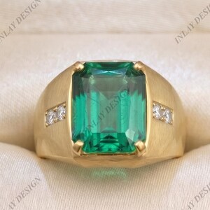 May include: A gold ring featuring a large, rectangular, emerald-cut green gemstone set in the center. The band has a brushed matte finish and includes two small, clear, round-cut accent stones on each side of the central green gemstone.