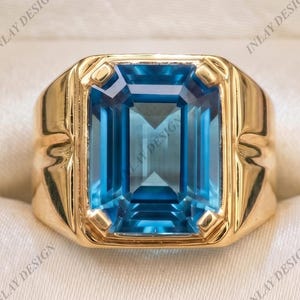 May include: A gold ring featuring a large, rectangular, emerald-cut blue gemstone set in a polished gold band. The ring is displayed inside a black jewelry box with a cream-colored interior lining. The text INLAY DESIGN is visible on the lining.