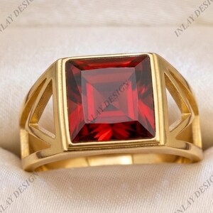 May include: A gold ring featuring a large, square-cut, deep red gemstone set in the center. The band has a geometric design with triangular cutouts on both sides. The ring is presented in a black box with a cream-colored interior lining.