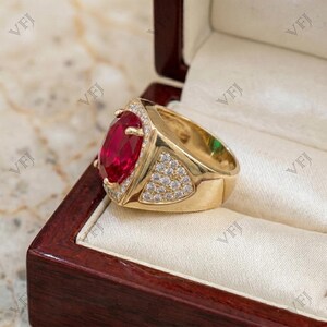 May include: A gold ring featuring a large, oval-cut, vibrant red gemstone set in the center. The band is accented with numerous small, clear, round-cut stones embedded along the sides of the setting, creating a bright, reflective surface.